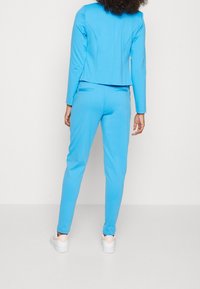 Blå figursydd jacka med sömdetaljer som matchas med smala byxor. Materialet verkar mjukt med en slät yta. Vita sneakers fulländar looken.