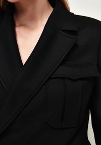 Černý blazer se strukturovaným designem, širokými klopami a dvojitými náprsními kapsami. Texturovaná tkanina s viditelnými švy.