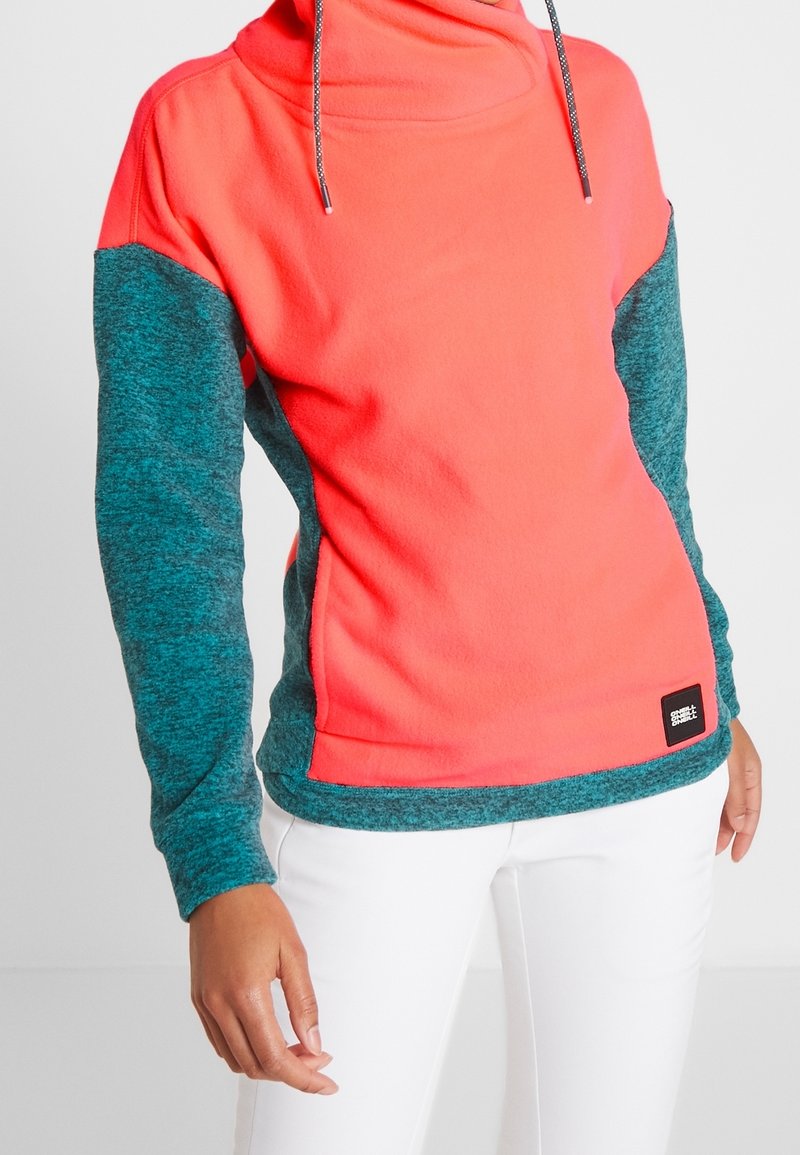 Sudadera de forro polar en coral brillante con mangas en teal. Presenta un cuello alto, cordón ajustable y una pequeña etiqueta de logo en la parte inferior izquierda.