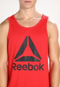 Débardeur rouge sans manches en tissu à texture lisse, avec un grand logo noir de Reebok au centre et un motif en triangle.