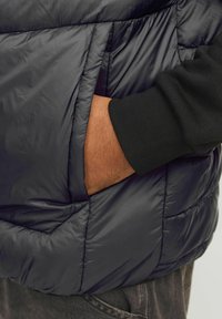 Dunkler Steppweste mit gepolsterter Textur, ausgestattet mit einer Seitentasche. Sichtbarer schwarzer Pulloverkopf darunter und eine Hand, die in die Tasche gesteckt ist.