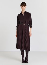 Woman wearing a dark brown satin buttoned shirt, pleated midi skirt with black belt, and knee-high dark brown boots, standing against white background.