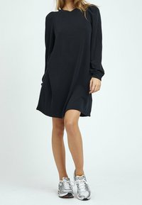 Svart långärmad klänning med avslappnad passform, med rund halsringning och draperade axeldetaljer. Kombinerad med silverfärgade träningsskor.