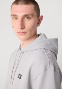 Lichtgrijze hoodie met een trekkoordcapuchon, een voorkant zak en een klein zwart label op de borst. Gladde textuur met zichtbare stikdetails.