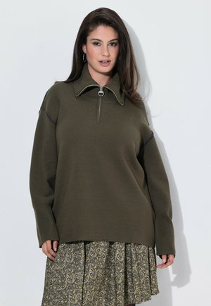 Vrouw met lang bruin haar, een donkergroen vest met rits en een patroonrijke olijfgroene rok, staand tegen een witte achtergrond.
