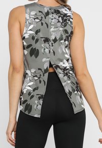 Bezrekawny top treningowy w kolorze szarym z kwiatowym wzorem; posiada rozcięcie na plecach i kontrastujące logo na górnej części pleców. Gładka faktura tkaniny.