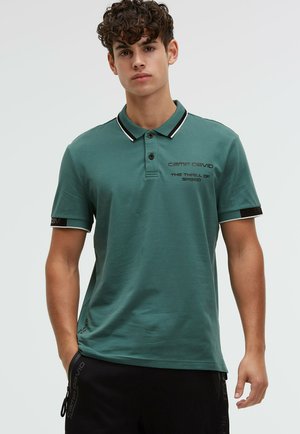 Junger Mann mit lockigem Haar, der ein grünes Poloshirt mit schwarzem und weißem Besatz und schwarze Shorts trägt, vor einem schlichten Hintergrund stehend.