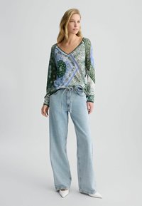 Kleurige blouse met lange mouwen en een V-hals, met een mix van groene, blauwe en witte patronen. Gecombineerd met lichtblauwe wijde jeans en witte hakken.