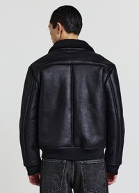 Uomo che indossa una giacca nera con colletto in pelliccia e jeans scuri, visto di spalle su uno sfondo chiaro e semplice.