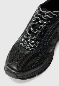 Chaussure de sport noire avec une tige en maille, des superpositions texturées et des lacets à motif noir et blanc, dotée d'un col rembourré et d'une semelle en caoutchouc.