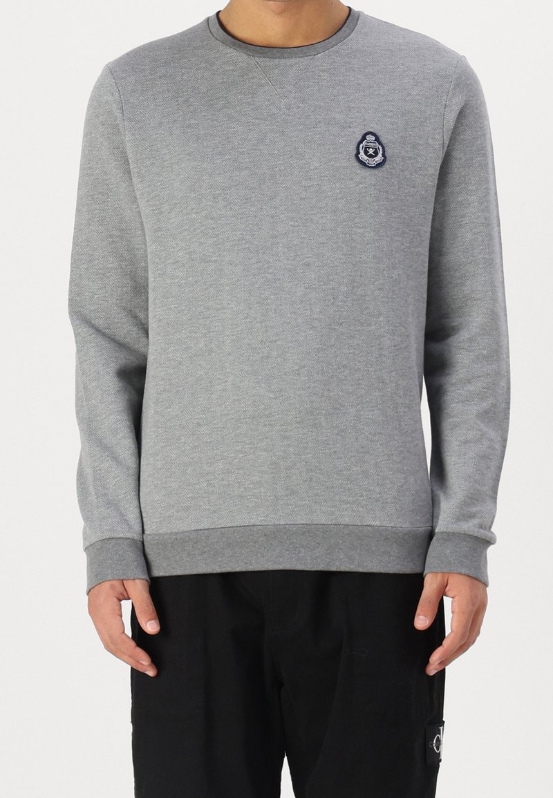 Homme portant un sweat-shirt gris clair à manches longues et col rond, avec un petit emblème brodé sur la poitrine gauche, associé à un pantalon noir.