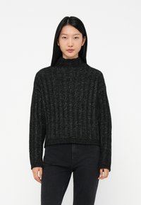 Jonge vrouw met lang zwart haar, gekleed in een zwarte ribgebreide hoge hals trui en hoogwijkende zwarte jeans, staand tegen een witte achtergrond.
