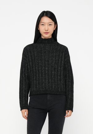 Giovane donna con lunghi capelli neri che indossa un maglione nero a coste con collo alto e jeans a vita alta neri, in piedi contro uno sfondo bianco.