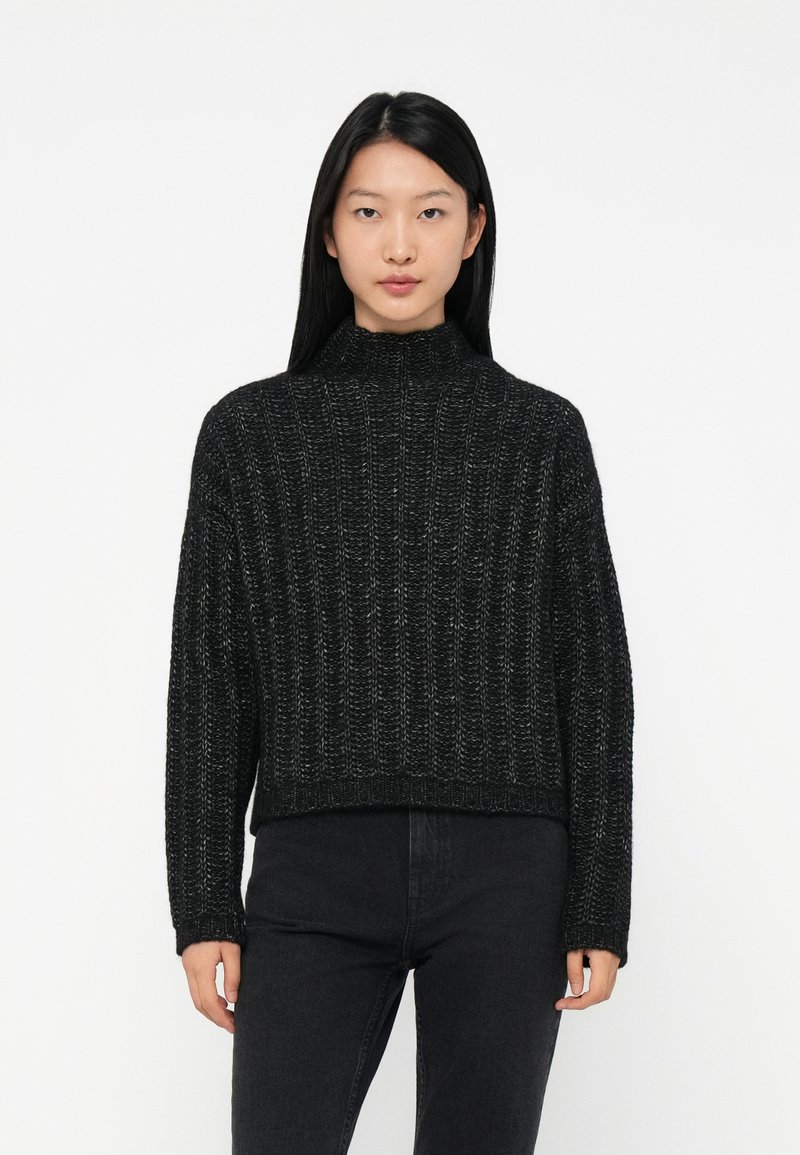 Jonge vrouw met lang zwart haar, gekleed in een zwarte ribgebreide hoge hals trui en hoogwijkende zwarte jeans, staand tegen een witte achtergrond.