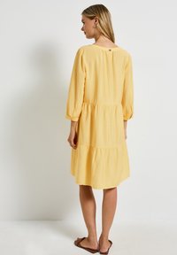 Žluté šaty do poloviny stehen s třičtvrtinovými rukávy, s vrstveným designem, vyrobené z lehké látky a s detailem knoflíku na zádech. Na nohou jsou ploché sandály.