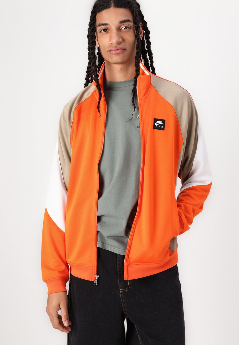 Orangene Trainingsjacke mit beigen, weißen und schwarzen Akzenten; mit durchgehendem Reißverschluss; über einem grauen Hemd getragen; zeichnet sich durch ein sportliches Design und eine glatte Textur aus.