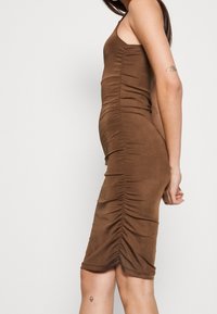 Hnědé přiléhavé šaty bez rukávů s nařasením podél boku a hladkým povrchem. Délka do poloviny lýtek.
