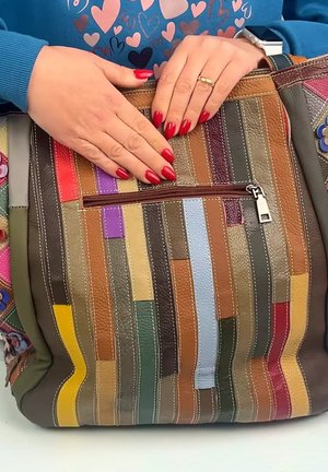 Hands with red nails on a multicolored striped leather bag with a zipper pocket, wearing a blue sweater with heart patterns.
