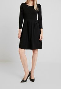 Svart långärmad klänning med rund halsringning, figurnära bodice och utsvängd kjol, tillverkad av ett slätt tyg. Bärs med svarta högklackade skor.