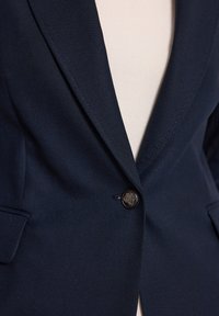 Granatowa marynarka z teksturowanej tkaniny, z pojedynczym dużym czarnym guzikiem, klapami w kształcie notch oraz bocznymi kieszeniami. Dopasowany krój z delikatnym połyskiem.