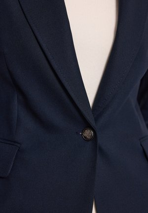 Granatowa marynarka z teksturowanej tkaniny, z pojedynczym dużym czarnym guzikiem, klapami w kształcie notch oraz bocznymi kieszeniami. Dopasowany krój z delikatnym połyskiem.