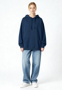 Donna in piedi che indossa una felpa oversize blu scuro, jeans a gamba larga azzurri e sneakers bianche su uno sfondo uniforme.