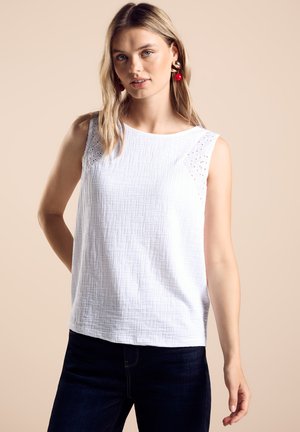 Witte mouwloze top met een gestructureerd oppervlak en oogjesaccenten op de schouders, gecombineerd met donkerblauwe jeans. Eenvoudig, casual ontwerp.