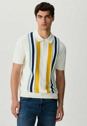 Junger Mann trägt ein weißes Poloshirt mit vertikalen gelben, marineblauen und hellblauen Streifen, kombiniert mit blauen Jeans, und steht vor einem schlichten Hintergrund.