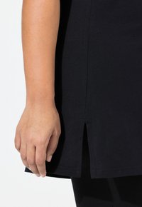 Černé, krátké bavlněné tričko s rozparkem na boku, které se vyznačuje hladkou texturou a jednoduchým designem. Na straně je vidět ruka.