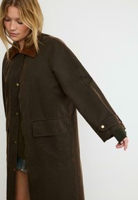 Cappotto lungo marrone con colletto in velluto a coste, bottoni a pressione e tasca frontale. Realizzato in un tessuto testurizzato e resistente con una vestibilità comoda.