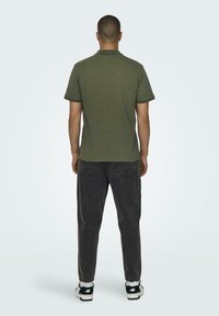 Olivgrünes T-Shirt mit kurzen Ärmeln, entspannter Passform, strukturiertem Stoff und subtilem Muster, kombiniert mit dunkelgrauen Jeans und Sneakers.