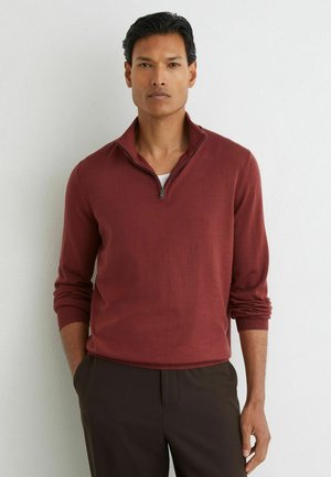 Mand iført en langærmet vinrød halv-lynlås sweater og mørke bukser står mod en hvid væg med hænderne i lommerne.