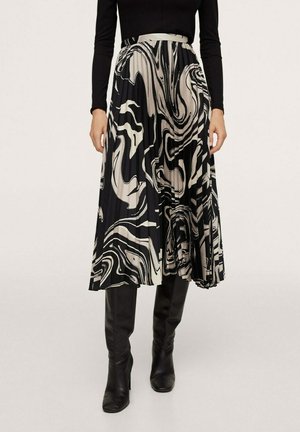 Kobieta w plisowanej spódnicy midi z abstrakcyjnym wzorem wirów w kolorach czarnym, białym i beżowym, w połączeniu z czarnym topem z długim rękawem i czarnymi kozakami za kolano.