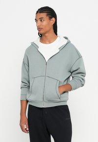 Svijetlozelena zip-up majica s rebrastom teksturom, bijelim unutarnjim podstavom i prednjim džepovima. Nosi se preko bijele majice s ležernim krojem.