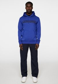 Felpa blu con tasca frontale e stampa nera del logo, abbinata a pantaloni blu navy e sneakers bianche. Tessuto morbido con vestibilità rilassata.