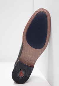 Semelle extérieure noire et marron avec motifs texturés et détails du logo. Forme arrondie avec matériau flexible et sections de couleurs contrastantes.