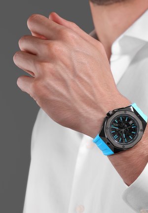 Mies, joka käyttää mustaa kelloa, jossa on sininen ranneke ja sinisiä yksityiskohtia kellotaulussa, säätelee valkoisen pitkähihainen paidan kalvosinta.