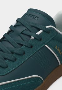Σκούρο πράσινο sneaker ARKK με πάνελ από σουέντ και δέρμα, κορδόνια στο ίδιο χρώμα, λευκή λεπτομέρεια και καφέ λαστιχένια σόλα σε κοντινή λήψη.