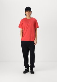 Tricou roșu din bumbac cu un mic logo alb pe piept, purtat cu pantaloni de trening negri și adidași negri. Croială simplă, lejeră.