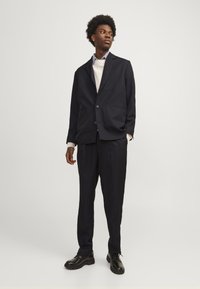 Abito blu navy con un blazer strutturato e pantaloni a sigaretta. Indossato sopra un maglione leggero, completato da scarpe stringate nere. Finitura in tessuto liscio.
