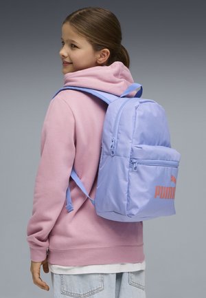 Niña joven con una sudadera rosa con capucha y una mochila Puma azul claro, sonriendo y mirando hacia un lado contra un fondo gris.