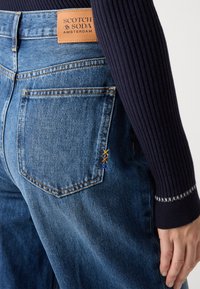 Denimjeans i mellomblått med et lærmerke. Funksjoner inkluderer to baklommer, kontraststing og dekorativ brodering i gult og blått.