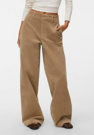 Personne portant un pantalon en velours côtelé beige taille haute et à jambes larges avec poches avant, des chaussures tissées foncées sans lacets, et un haut blanc court à manches longues visible.
