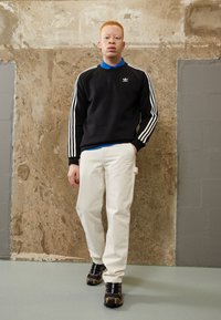 Un sweat noir avec des rayures blanches, une chemise col bleu en dessous, un pantalon crème, des baskets noires avec des accents orange, se tenant devant un fond texturé.