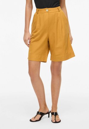 Femme portant un short taille haute jaune moutarde ajusté avec fermeture par boutons, associé à des sandales noires à talons et un haut noir.