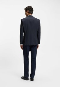 Abito blu navy con vestibilità su misura, giacca monopetto e pantaloni coordinati. Tessuto liscio e dettagli sottili sui polsini delle maniche. Scarpe nere.