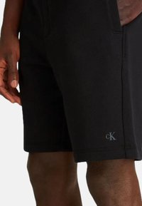 Pantaloni corti neri realizzati in materiale morbido, con una vestibilità slim, tasche laterali e un piccolo logo vicino all'orlo. Texture liscia con linee pulite.