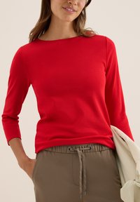 Haut rouge à manches longues avec un décolleté bateau, en tissu doux, associé à un pantalon beige à taille élastique avec un détail de cordon.
