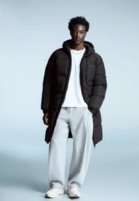 Geacă puf neagră cu glugă, închidere cu fermoar și buzunare laterale, purtată peste un tricou alb și pantaloni de trening gri, completată de adidași albi.