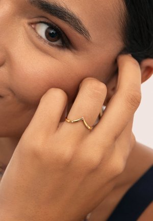 Anillo de oro con un diseño ondulado, que presenta pequeñas piedras preciosas de colores. La imagen muestra el anillo en un dedo, con una mano descansando cerca de la cara.